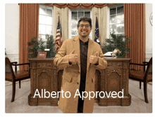 a man in a tan coat is giving a thumbs up in front of a desk that says alberto approved on it