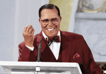 a man in a red suit and bow tie stands at a podium giving a speech