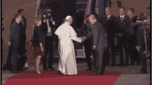 a man in a suit is shaking hands with a pope on a red carpet in front of a group of people .