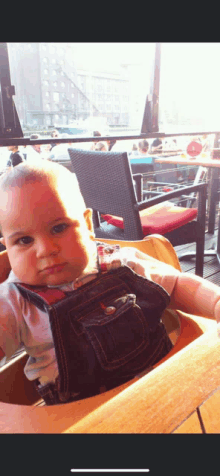 a baby in overalls sits in a high chair looking at the camera