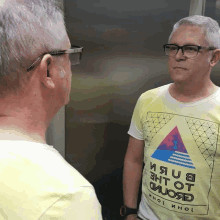 a man wearing glasses and a yellow t-shirt that says " ияu8 " on it looks at his reflection