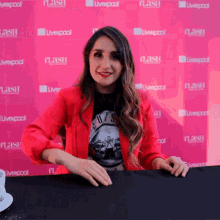 a woman in a red jacket is sitting at a table in front of a pink wall that says ' liverpool ' on it