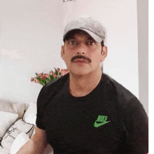 a man with a mustache wearing a hat and a black nike shirt is sitting on a couch in a living room .