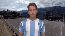 a man wearing a blue and white striped shirt stands in front of a soccer field with mountains in the background