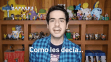 a man in a plaid shirt says " como les decia " in front of a shelf full of stuffed animals