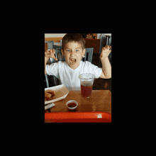 a young boy sitting at a table with a cup of soda and a container of ketchup