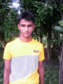 a young man wearing a yellow and white striped shirt stands in front of a tree