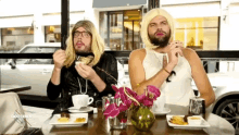 two men are sitting at a table with plates of food and a vase of flowers with the word goodies on the bottom