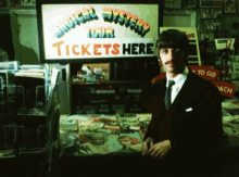 a man in a suit and tie stands in front of a sign that says magical mystery tour tickets here