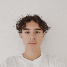 a young man with curly hair is wearing a white t-shirt and looking at the camera .