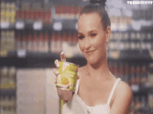 a woman in a white tank top is holding a can of food in front of a grocery store shelf