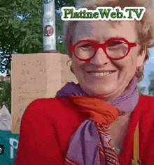 a woman wearing red glasses and a purple scarf smiles in front of a sign that says platineweb tv