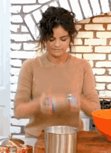 a woman in a tan sweater is standing in a kitchen with a bowl and a brick wall behind her .