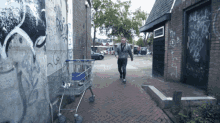 a man walking down a sidewalk with a shopping cart in front of a wall that has graffiti on it