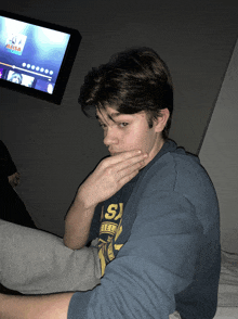 a young man covering his mouth with his hand while wearing a blue shirt with the letter s on it