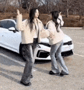 two young women are standing next to each other in front of a white car in a parking lot .