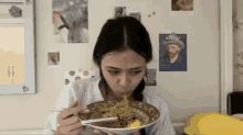 a woman is eating a bowl of food with chopsticks in front of a wall with posters of vincent van gogh on it