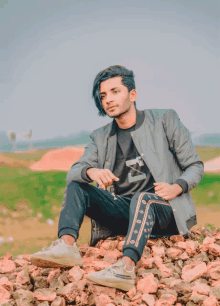 a young man sits on a pile of rocks wearing a gray jacket and a black shirt with the letter m on it