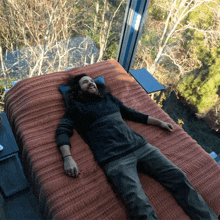 a man with long hair is laying on a bed in front of a window with trees in the background