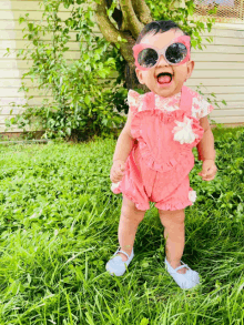 a baby girl wearing pink overalls and sunglasses stands in the grass