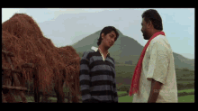 a man in a striped shirt stands next to another man in a white shirt in front of a pile of hay in a field