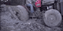 a man is driving a tractor through a muddy field with a red towel on the back of it .