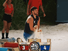 a man in a black tank top is kneeling in the sand near a scoreboard with the number 3 and 4 on it