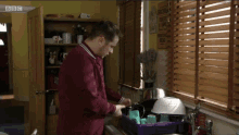 a man is washing dishes in front of a window with blinds and a bbc logo on the corner of the screen