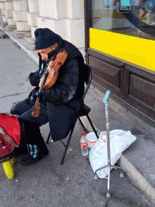 a man sits on a chair playing a violin in front of a store with a bag that says aquafina on the sidewalk