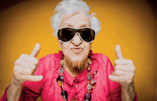 an elderly woman wearing sunglasses and a pink shirt gives a thumbs up