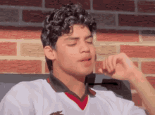 a young man with curly hair wearing a white shirt with a red collar is sitting in front of a brick wall .