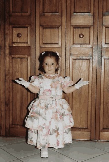 a little girl wearing a floral dress and white gloves stands in front of a wooden wall