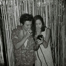 a black and white photo of a man singing into a microphone next to a woman holding a cell phone .