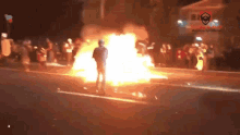 a man stands in front of a large fire with a shield on the bottom right corner of the image