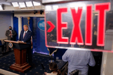 a man stands at a podium behind a red exit sign