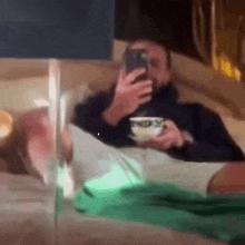 a man is laying on a couch holding a cup of coffee and looking at his cell phone .