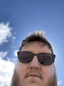 a man wearing sunglasses looks at the camera with a blue sky behind him