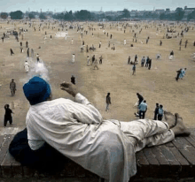 a man is laying on a wooden ledge smoking a cigarette and looking at a cricket field .