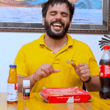 a man in a yellow shirt is sitting at a table with a box of pizza and a bottle of coke