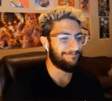 a man with a beard and glasses is sitting in a chair in front of a wall with posters on it .