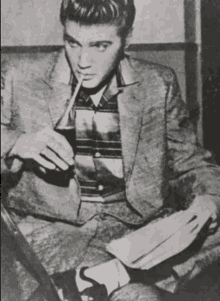 elvis presley is drinking a soda through a straw while sitting on a chair .