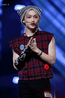 a young man wearing a plaid shirt and a beanie stands in front of a microphone on a stage .