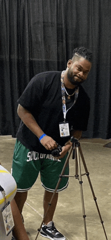 a man in a black shirt and green shorts is standing next to a tripod with a camera