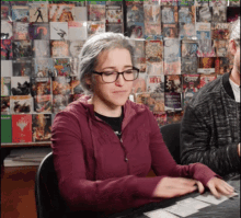 a woman wearing glasses sits at a table in front of a wall of comic books including one called spiderman