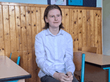 a young man in a white shirt sits at a desk with his hands folded