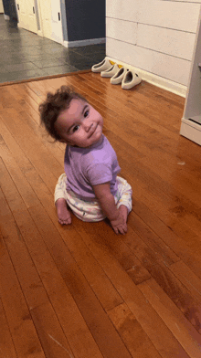 a baby in a purple shirt sits on a wooden floor