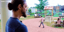 a man with a ponytail and a beard is looking at a group of people sitting at picnic tables ..