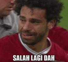 a soccer player is crying on the field while wearing a red shirt and a caption that says salah lagi dah .