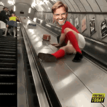 a man in red socks and a red shirt is sliding down an escalator with a football today advertisement behind him