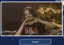 a woman making a heart with her hands in front of a christmas tree and a button that says " tolyan "